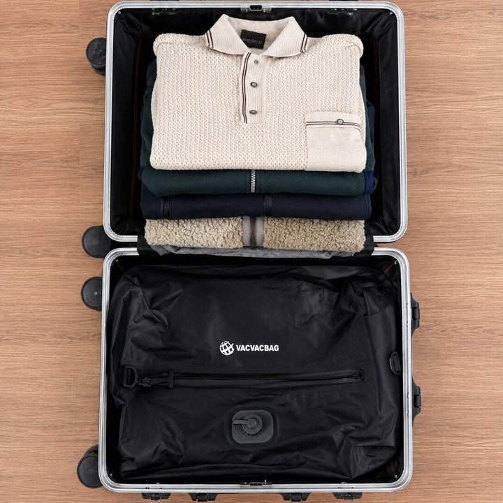 An open silver suitcase on a wood floor, showing a neatly folded cream knit polo shirt and other folded garments inside, with the black Vacvacbag compression bag in the bottom compartment.
